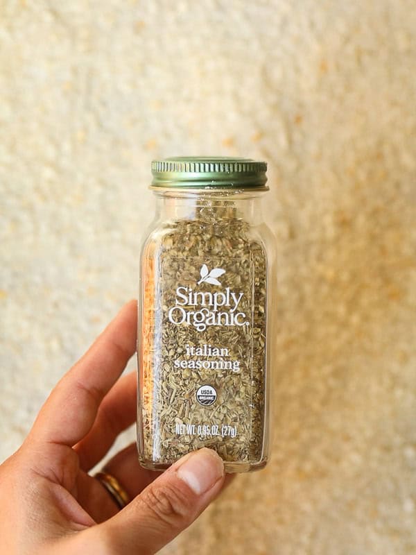 Hand holding a jar of Simply Organic Italian Seasoning with a metal green lid. Background is a textured, light-colored surface.